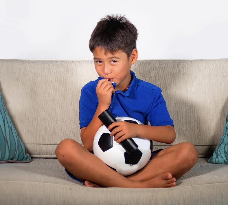 Een kind zit op de bank met een bal en de afstandsbediening van de tv